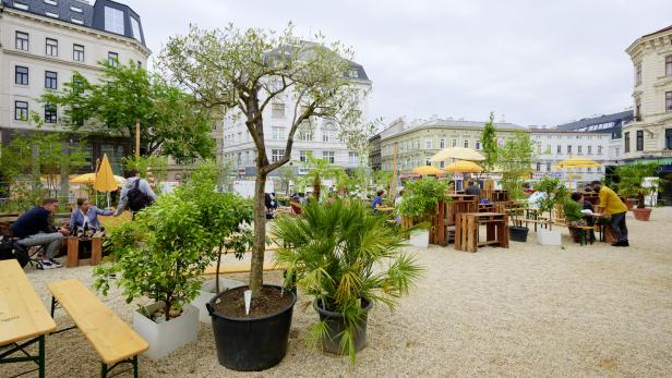 Ein belebter Außenbereich mit Tischen und Sonnenschirmen in einer städtischen Umgebung.