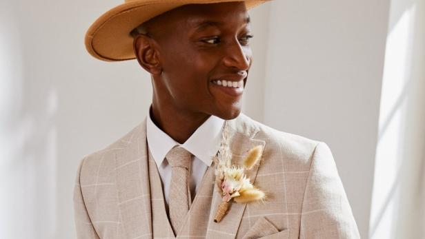 Ein lächelnder Mann mit Hut und beigefarbenem Anzug mit Blumenschmuck.