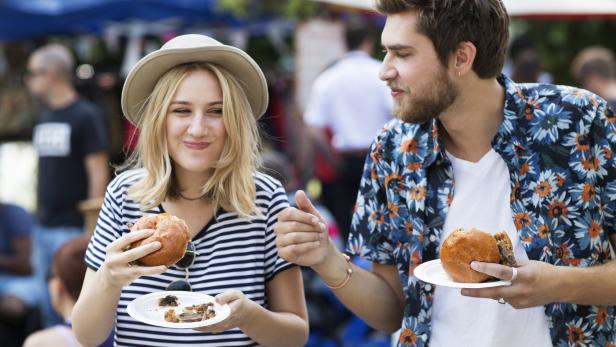 Eine Frau mit Hut und ein Mann essen Burger auf einem Markt.