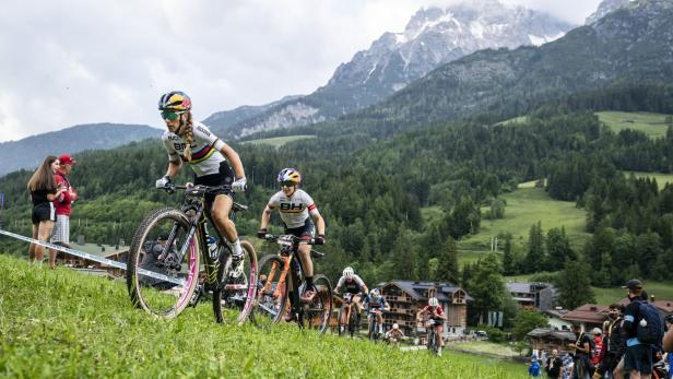 Mehrere Mountainbiker fahren einen grasbewachsenen Hügel hinauf, im Hintergrund Berge.