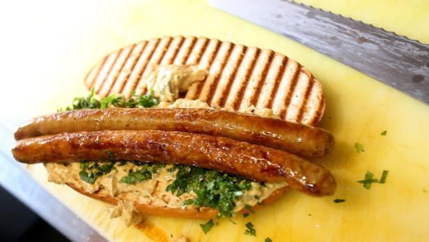 Ein Sandwich mit Würstchen, Kräutern und Soße auf einem gelben Schneidebrett.