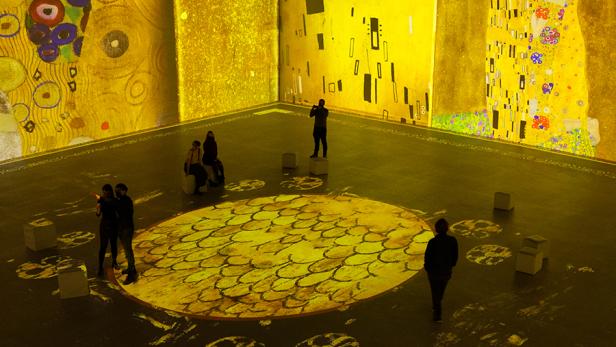 Besucher in einer Ausstellung, in der Werke von Gustav Klimt auf Wände und Boden projiziert werden.