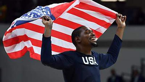 Ein lächelnder Mann mit einem T-Shirt mit der Aufschrift „USA“ hält eine amerikanische Flagge hoch.
