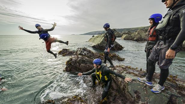 Eine Gruppe von Personen springt von Felsen ins Meer, bekleidet mit Neoprenanzügen und Helmen.