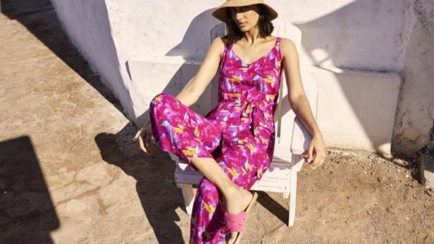 Eine Frau mit Hut und buntem Jumpsuit sitzt auf einem Stuhl im Freien.