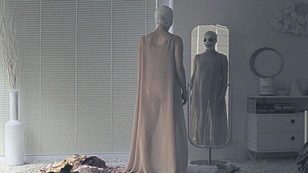 Eine in ein langes Kleid gehüllte Person mit bandagiertem Kopf steht vor einem Spiegel.