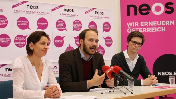 Drei Personen sitzen vor einem rosafarbenen Hintergrund mit dem Logo der Partei NEOS.