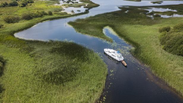 Ein weißes Boot fährt auf einem schmalen Flusslauf durch eine grüne Landschaft.