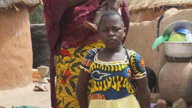 Eine Frau mit einem Baby auf dem Arm und ein Mädchen stehen vor traditionellen Hütten.