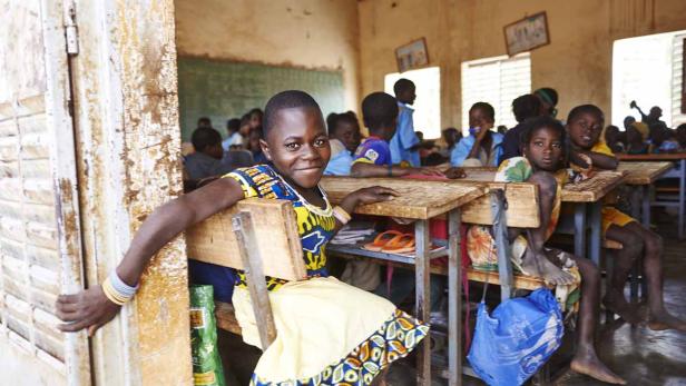 Ein Mädchen sitzt in einem Klassenzimmer und lächelt in die Kamera.