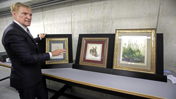 Ein Mann präsentiert drei gerahmte Kunstwerke mit Zeichnungen von Pflanzen und einem Hasen.