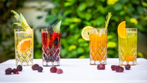 Vier Gläser mit bunten Cocktails, garniert mit Früchten und Kräutern, stehen auf einem Tisch.