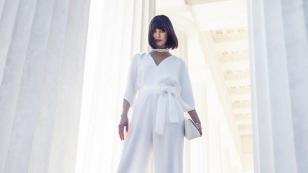 Eine Frau in einem weißen Hosenanzug posiert vor einem Säulengang.