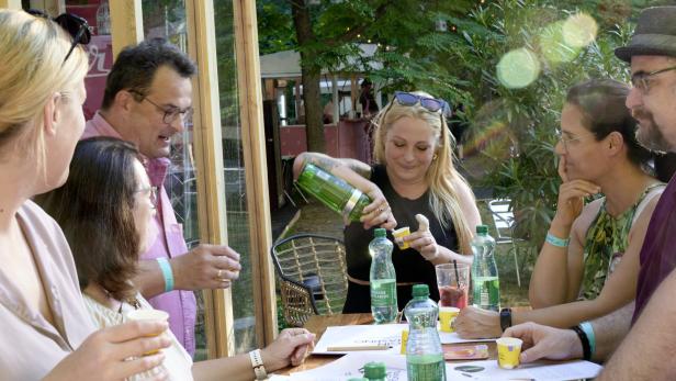 Eine Gruppe von Menschen sitzt an einem Tisch, während eine Frau Getränke aus einer grünen Flasche einschenkt.