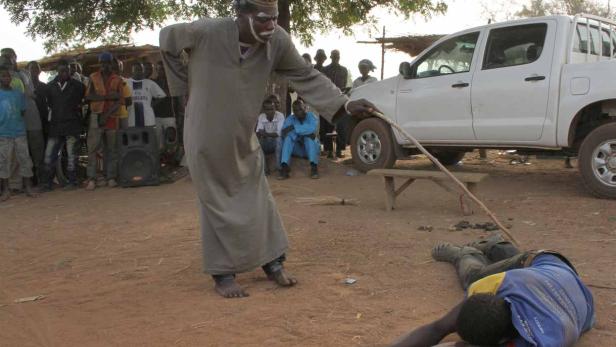 Ein Mann mit Gesichtsbemalung steht mit einem Stock über einer am Boden liegenden Person.