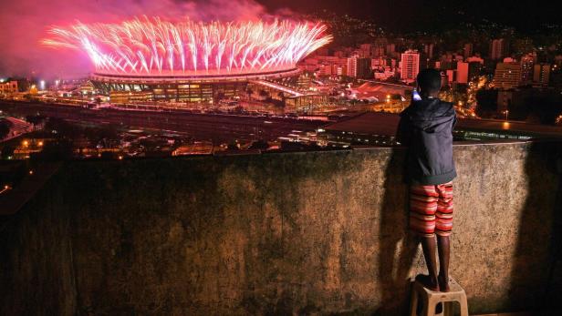 Ein Junge steht auf einem Hocker und beobachtet ein Feuerwerk über einem Stadion in der Stadt.