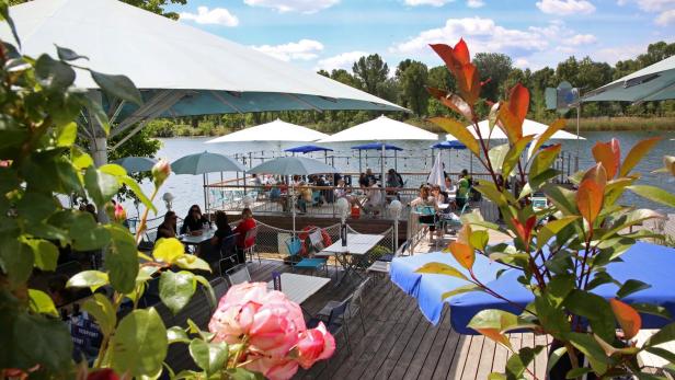 Ein Restaurant am See mit Gästen unter Sonnenschirmen auf einer Terrasse.