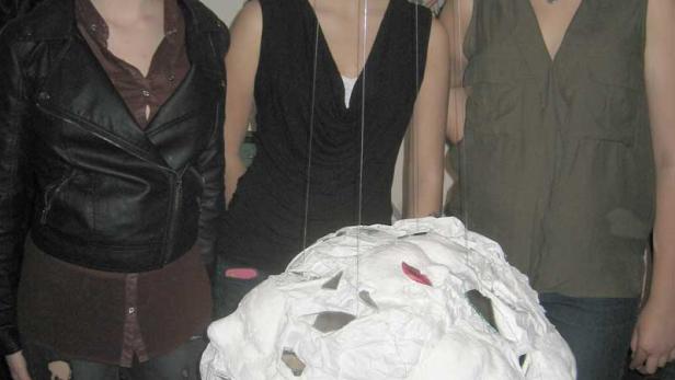 Drei junge Frauen stehen vor einer weißen, gesichtsförmigen Skulptur.