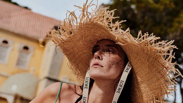 Eine Frau mit Strohhut, grünem Kleid und einer Tasche der Marke Loewe vor einem Gebäude.