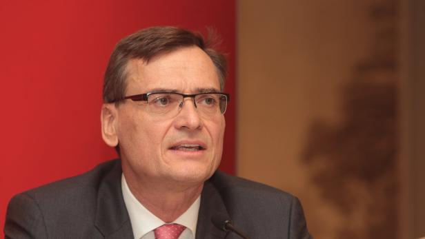 Ein Mann mit Brille und Anzug spricht vor einem roten Hintergrund.