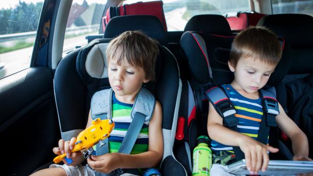 Zwei kleine Jungen in gestreiften T-Shirts sitzen in Kindersitzen auf der Rückbank eines Autos und spielen mit einem Spielzeugflugzeug und einem Tablet.