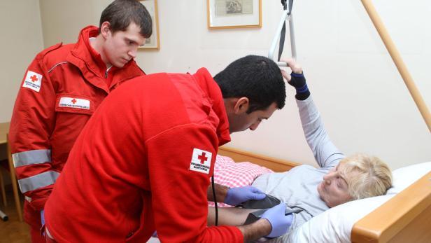 Zwei Sanitäter des Österreichischen Roten Kreuzes kümmern sich um eine ältere Patientin im Bett.