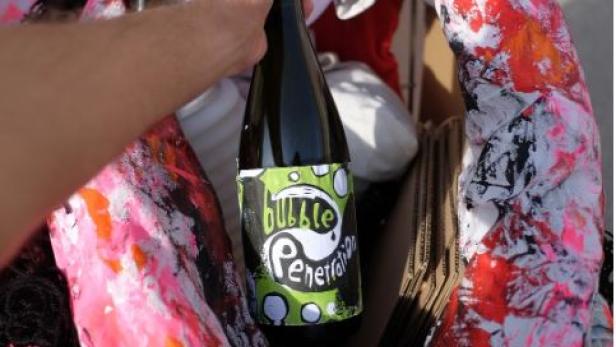 Eine Hand hält eine Flasche „Bubble Penetration“ in einem Karton mit einer bemalten Skulptur.