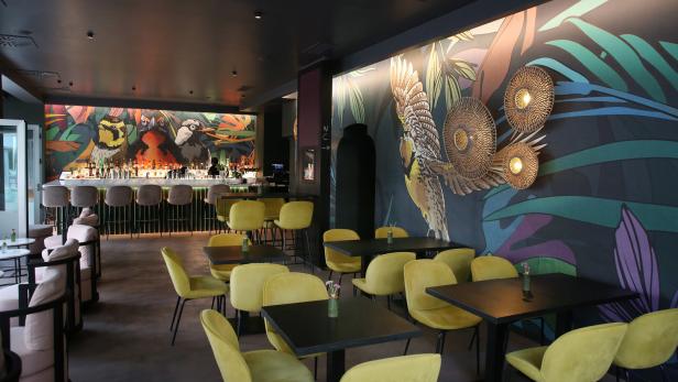 Innenansicht einer Bar mit Tischen, Stühlen und einer Wandmalerei mit Vogelmotiven.