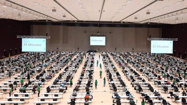 Große Halle mit vielen Menschen an Einzeltischen, die eine Prüfung ablegen, auf Leinwänden steht „29 Minuten“.