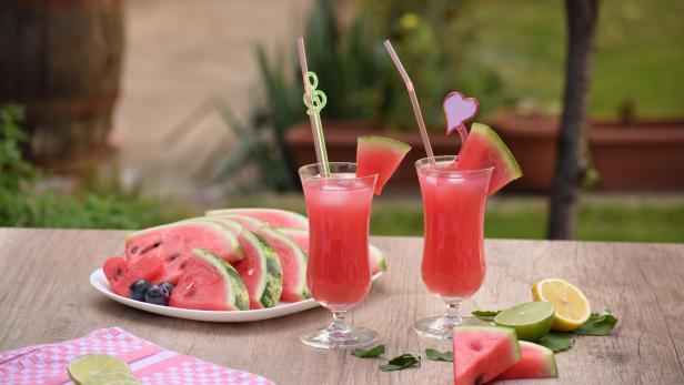 Zwei Gläser Wassermelonensaft stehen auf einem Holztisch, dekoriert mit Limetten und Melonenstücken.