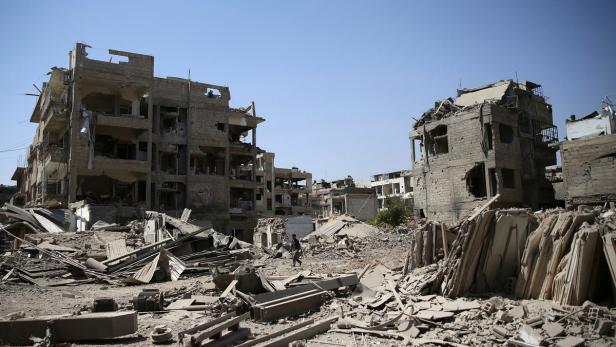 Zwei Männer gehen durch eine zerstörte Stadtlandschaft mit Trümmern und beschädigten Gebäuden.