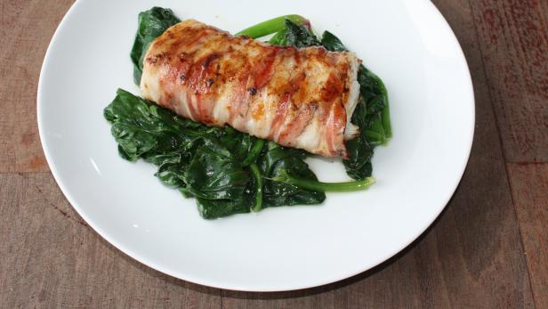 Ein mit Speck umwickeltes Fischfilet auf einem Bett aus Spinat auf einem weißen Teller.
