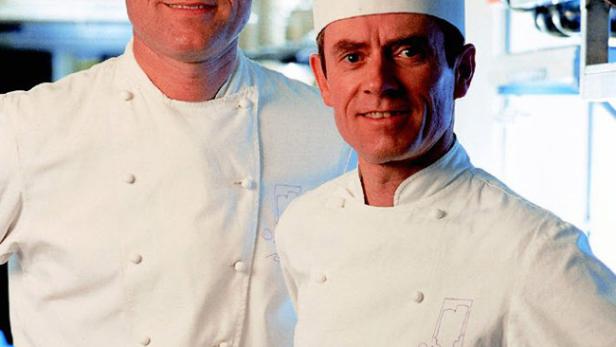 Zwei Köche mit Kochmützen und weißen Uniformen stehen in einer Küche.