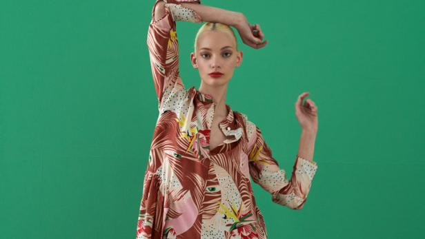 Eine Frau mit blonden Haaren posiert in einem gemusterten Kleid vor einem grünen Hintergrund.