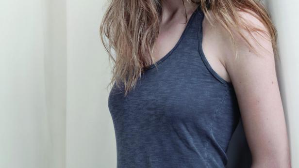 Eine Frau mit langen, gewellten Haaren und einem grauen Tanktop lehnt an einer Wand.