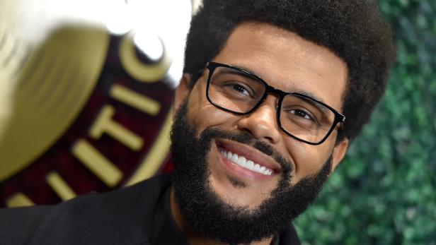 Der lächelnde Sänger The Weeknd mit Brille und Bart vor grünem Hintergrund.