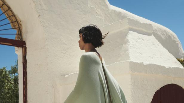 Eine Frau geht in einem hellgrünen Poncho mit Streifen vor einem weißen Gebäude entlang.