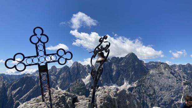 Gipfelkreuz und Glocke auf einem Berg mit Blick auf eine Bergkette.
