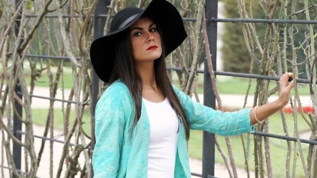 Eine Frau mit Hut und türkisfarbener Jacke posiert vor einem Rosengitter.