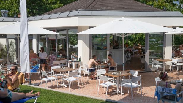 Gäste sitzen an Tischen im Freien vor einem Strandclub.