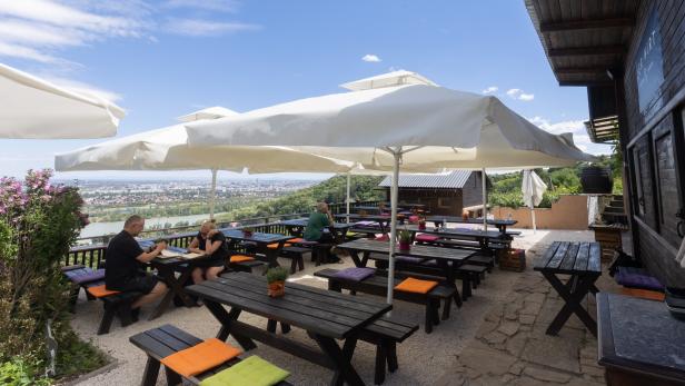 Ein Restaurant im Freien mit Tischen und Bänken unter Sonnenschirmen bietet einen Blick auf die Stadt.