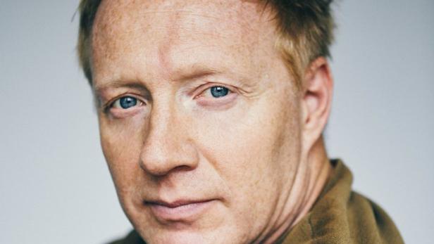 Porträt eines Mannes mit roten Haaren und blauen Augen, der einen braunen Pullover trägt.