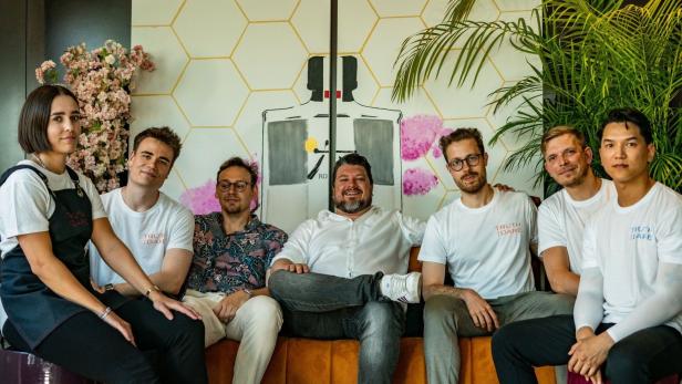 Eine Gruppe von sieben Personen sitzt auf einer Couch vor einem Wandbild.