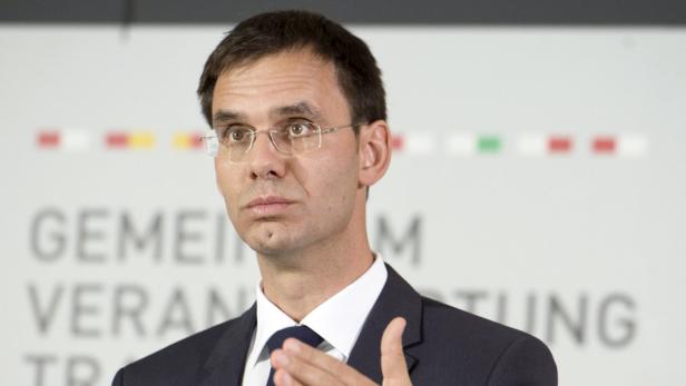 Ein Mann mit Brille und Anzug gestikuliert vor einem unscharfen Hintergrund.