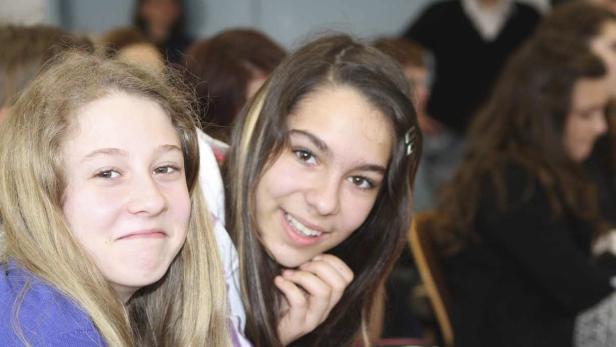 Zwei junge Frauen lächeln in die Kamera, im Hintergrund weitere Personen.