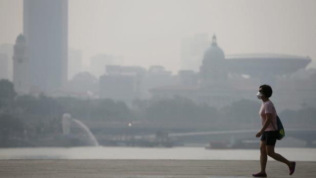 Eine Frau mit Maske geht vor der verschmutzten Skyline von Singapur entlang.