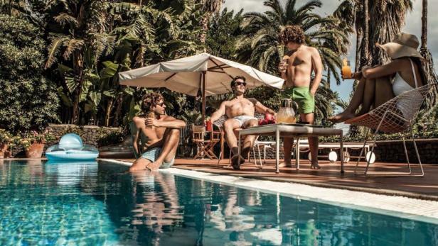 Eine Gruppe von Menschen entspannt sich mit Getränken an einem sonnigen Pool unter Palmen.