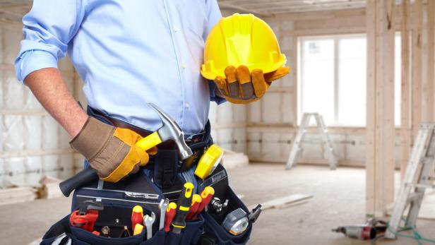 Ein Bauarbeiter hält einen gelben Schutzhelm in der Hand, mit Werkzeug am Gürtel vor einer Baustelle.