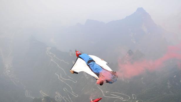 Ein Wingsuit-Pilot fliegt mit roter Rauchwolke vor einer Berglandschaft.