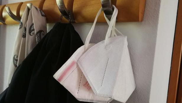 Zwei weiße FFP2-Masken hängen an einer Garderobe neben einem schwarzen Kleidungsstück und einem gemusterten Schal.